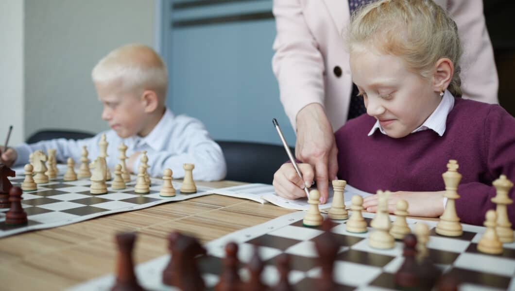 Smiling schoolgirl in purple sweater writing down chess moves while teacher pointing at notebook