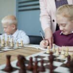Smiling schoolgirl in purple sweater writing down chess moves while teacher pointing at notebook