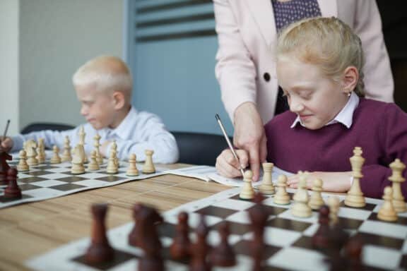 Smiling schoolgirl in purple sweater writing down chess moves while teacher pointing at notebook