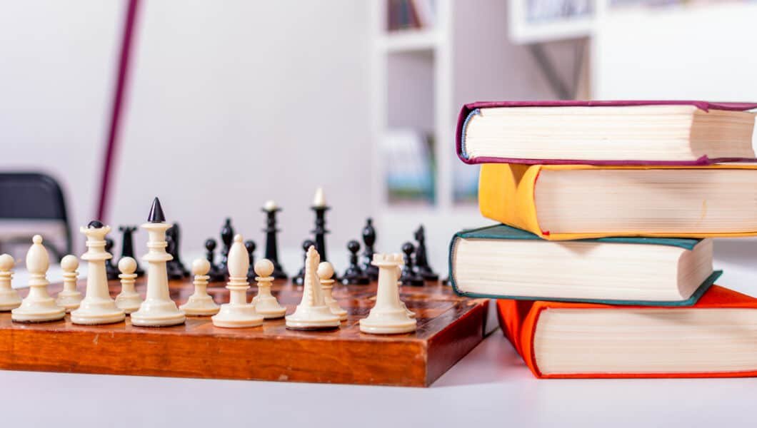 Wooden chessboard with white and black chess pieces arranged. Nearby lies stack of thick multi-colored books.