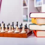 Wooden chessboard with white and black chess pieces arranged. Nearby lies stack of thick multi-colored books.