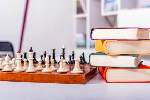 Wooden chessboard with white and black chess pieces arranged. Nearby lies stack of thick multi-colored books.